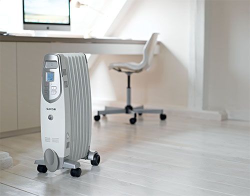 Un radiateur à bain d'huile blanc et gris, monté sur roulettes, se trouve dans un bureau au sol clair, à côté d'un bureau et d'une chaise.