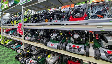 Des tondeuses à gazon exposées dans un magasin ; des modèles et des couleurs variés sur plusieurs étagères, éclairées par des lampes zénithales.