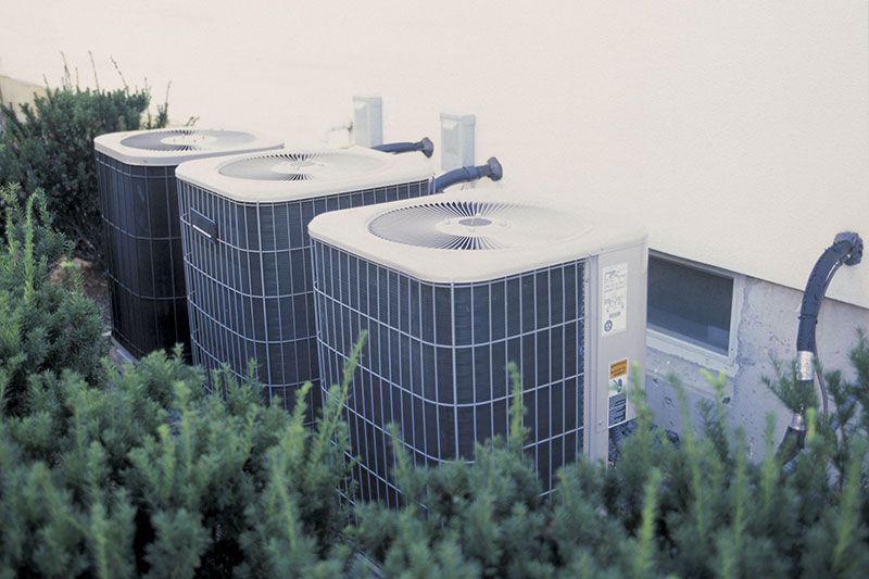 Trois unités de climatisation à l'extérieur d'un bâtiment, entourées de buissons verts.