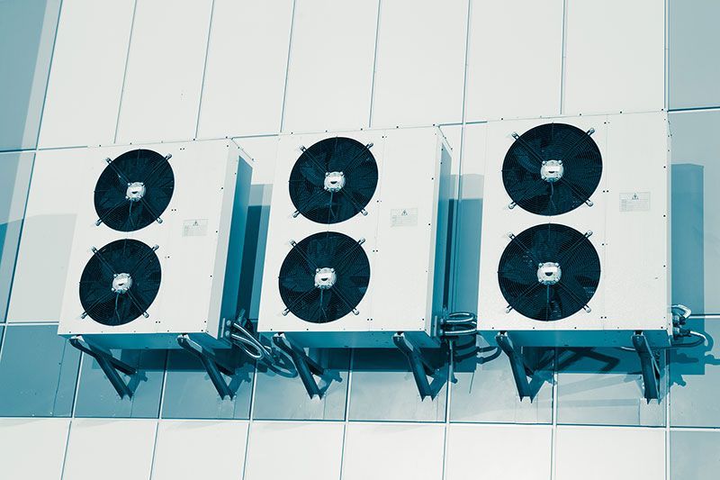 Trois unités de climatisation blanches fixées sur un mur carrelé, chacune dotée de deux pales de ventilateur noires.