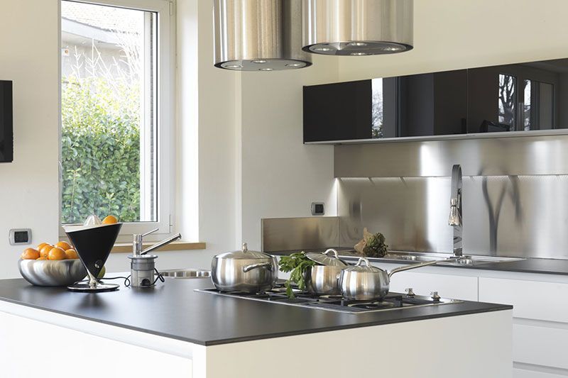 Îlot de cuisine moderne avec plaque de cuisson, casseroles, crédence en acier inoxydable, fenêtre et corbeille à fruits.