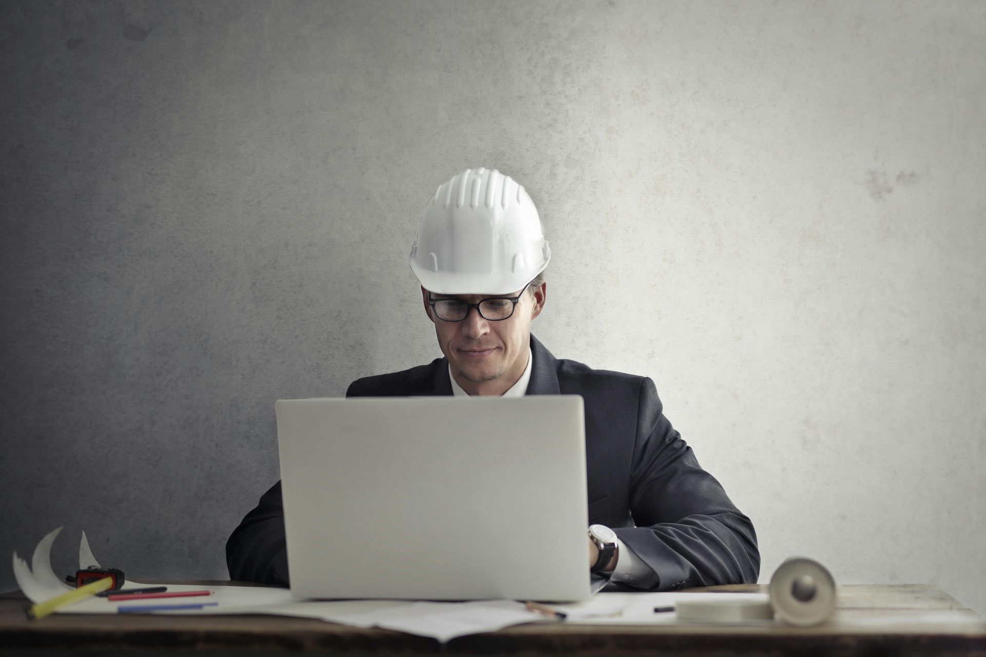 Ingeniero con casco trabajando en un portátil en un escritorio con papeles y un rollo de cinta adhesiva.