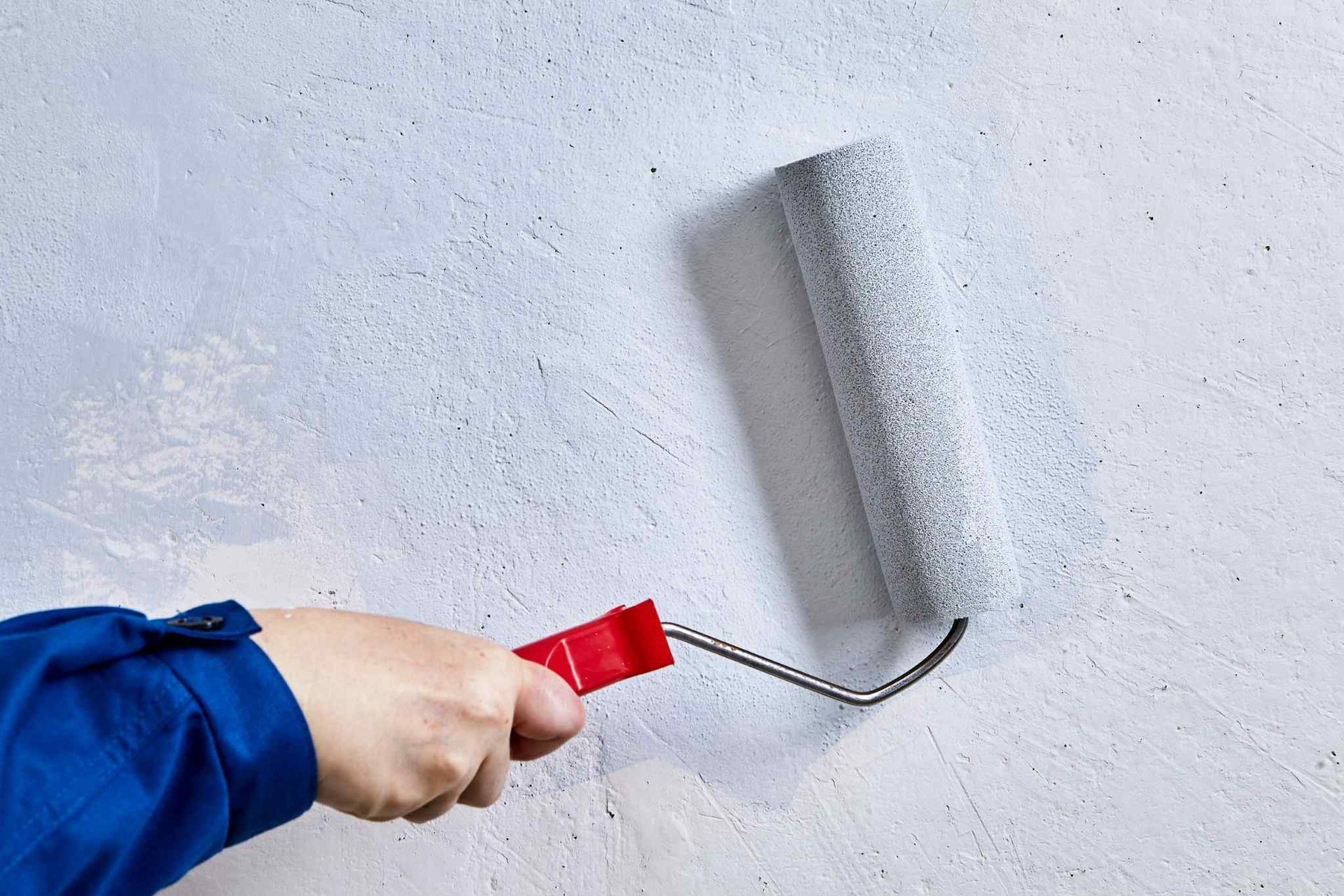 Mano sosteniendo un rodillo de pintura rojo pintando una pared gris de blanco.