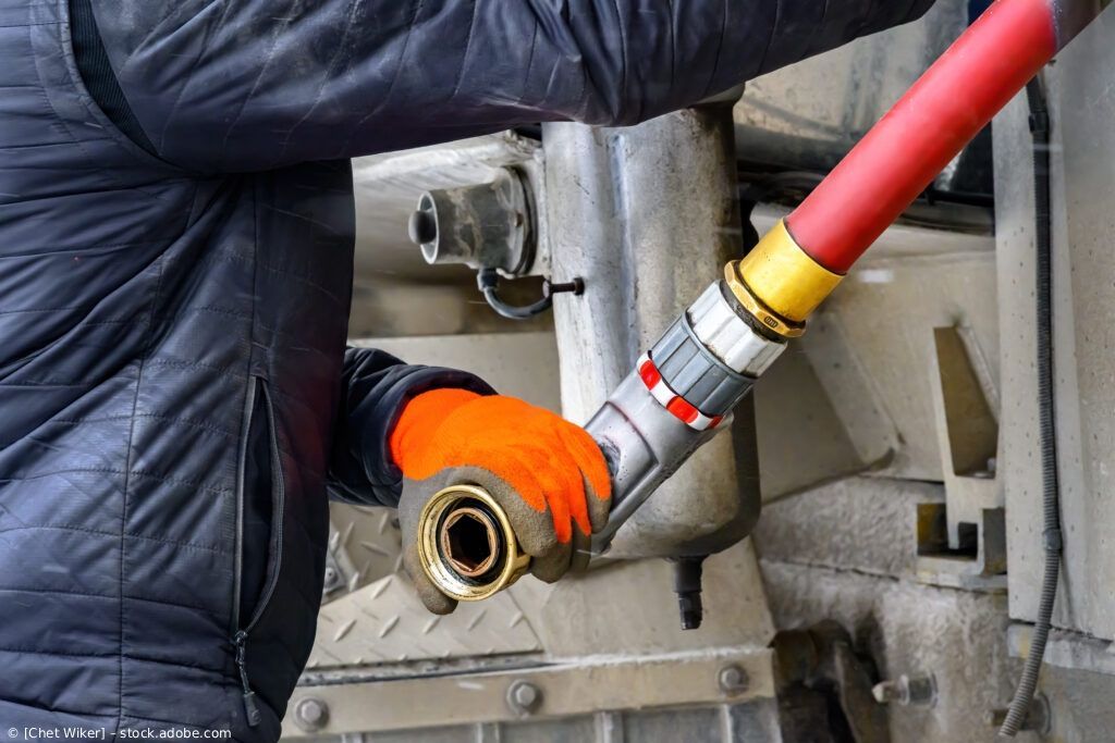 Ein Arbeiter mit orangefarbenen Handschuhen hält eine rote Schlauchdüse an einen metallischen Maschinenanschluss.