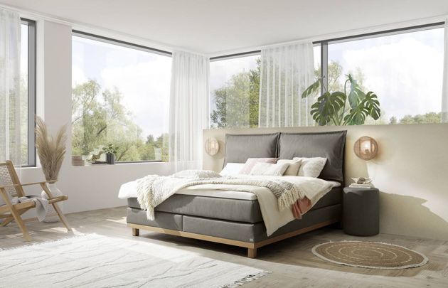 Bett mit weisser Bettwäsche und Kissen, hölzernes Kopfteil, in der Nähe eines Fensters mit Vorhängen.