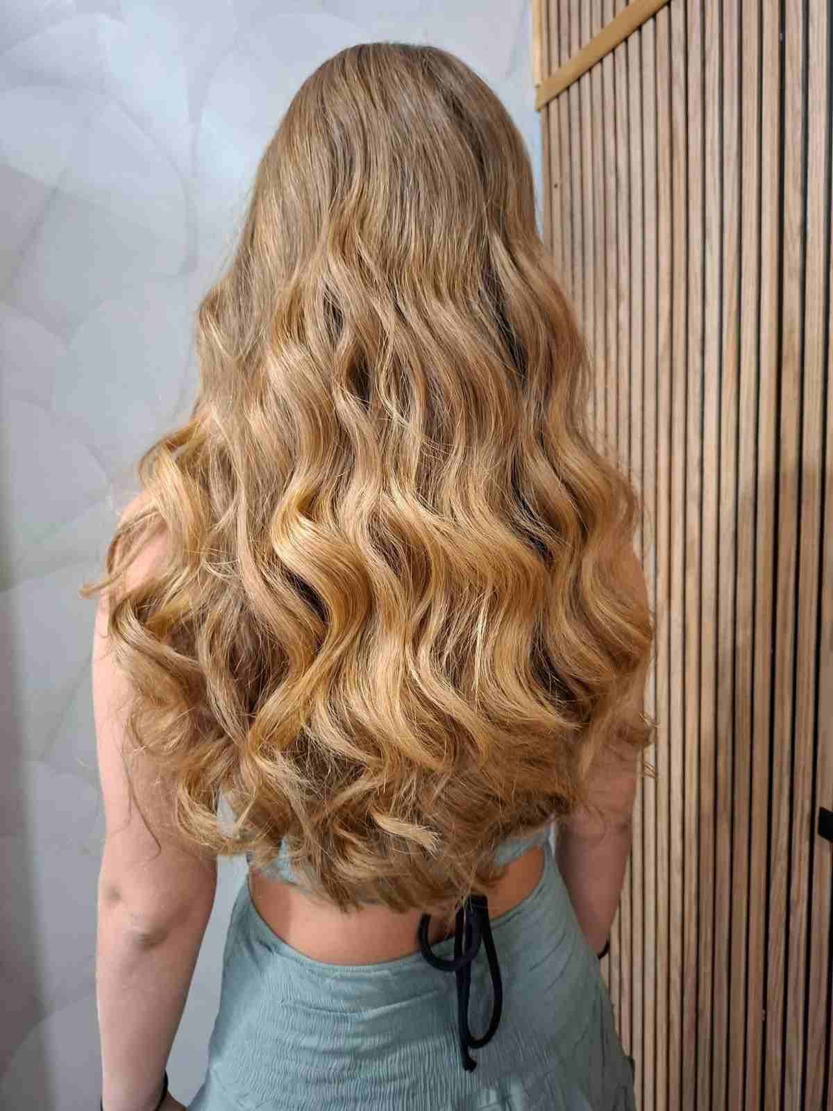 Una larga melena rubia y ondulada cae en cascada por la espalda de una persona con una blusa azul sin espalda.