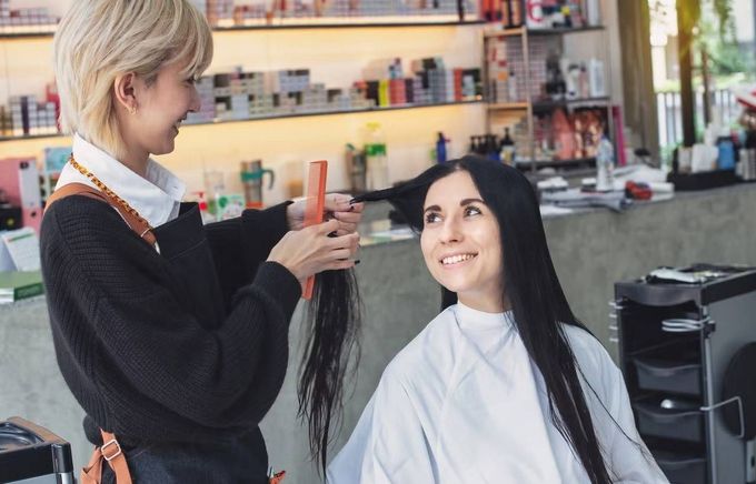 Peluquera arreglando el largo cabello negro de una clienta sentada en un salón de belleza.