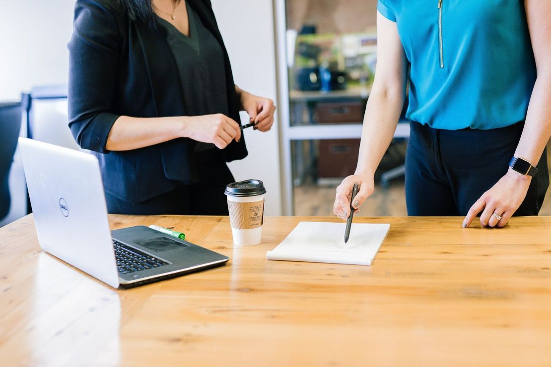 Deux personnes discutent de documents administratifs à une table en bois, un ordinateur portable et un café à la main, dans un bureau lumineux.