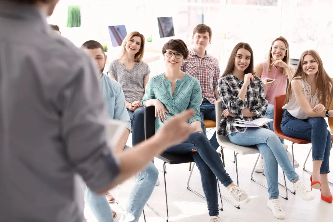 Un présentateur s'adresse à un public assis dans une salle de classe ou un atelier lumineux.