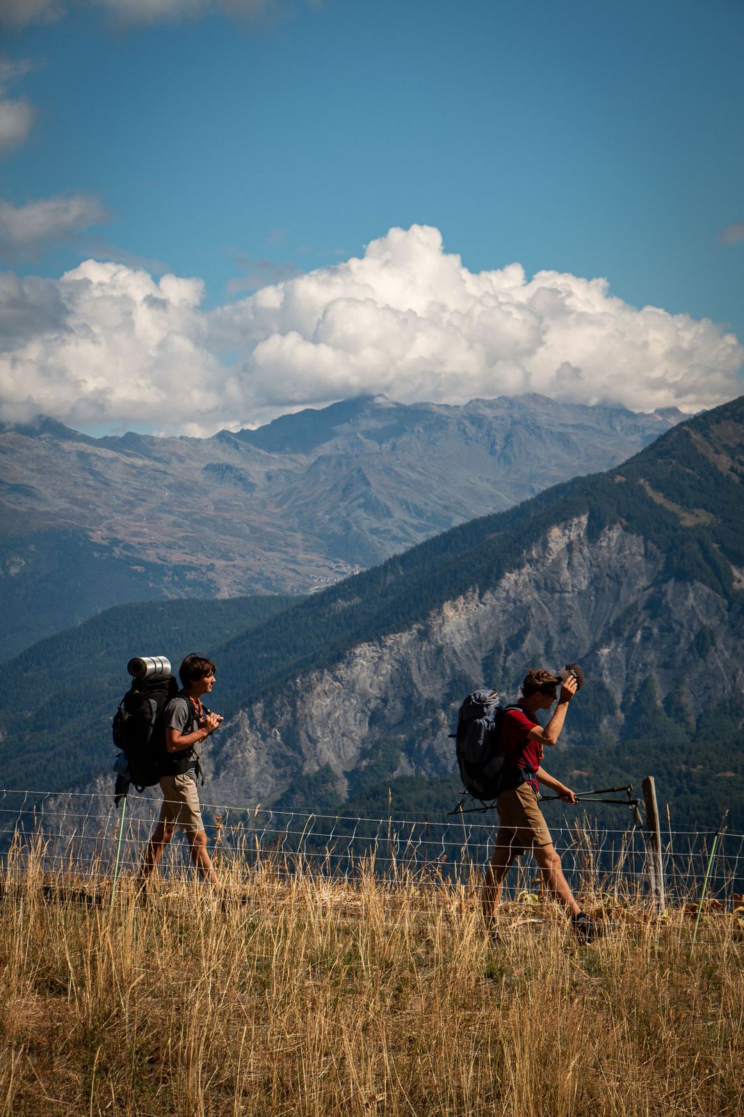 Deux randonneurs avec des sacs à dos marchent dans l'herbe sèche au pied des montagnes, sous un ciel bleu nuageux.