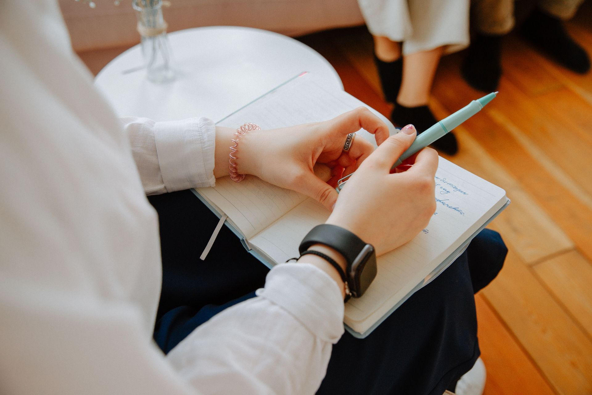 Une personne écrivant dans un carnet, assise sur une chaise blanche, tenant un stylo turquoise au-dessus d'un parquet.