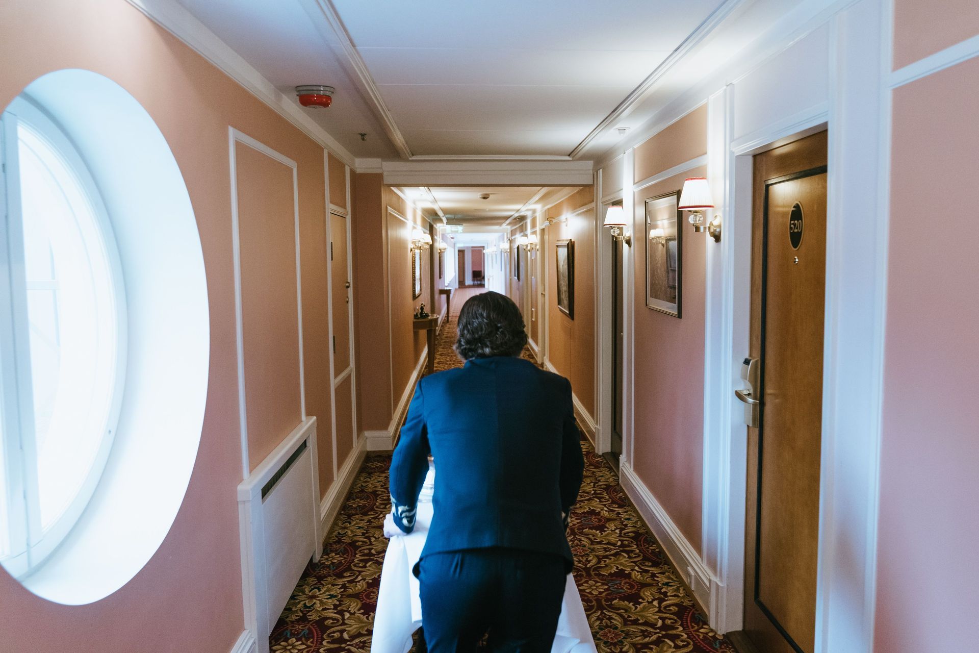 Une personne marche dans un couloir d'hôtel avec des fenêtres rondes, du sol recouvert de moquette et des appliques murales.