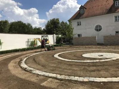 Ein Mann rollt Rasen in einem runden Beet. Im Hintergrund ein Gebäude mit rundem Fenster und eine Mauer mit Wandmalerei. Sonniger Tag.