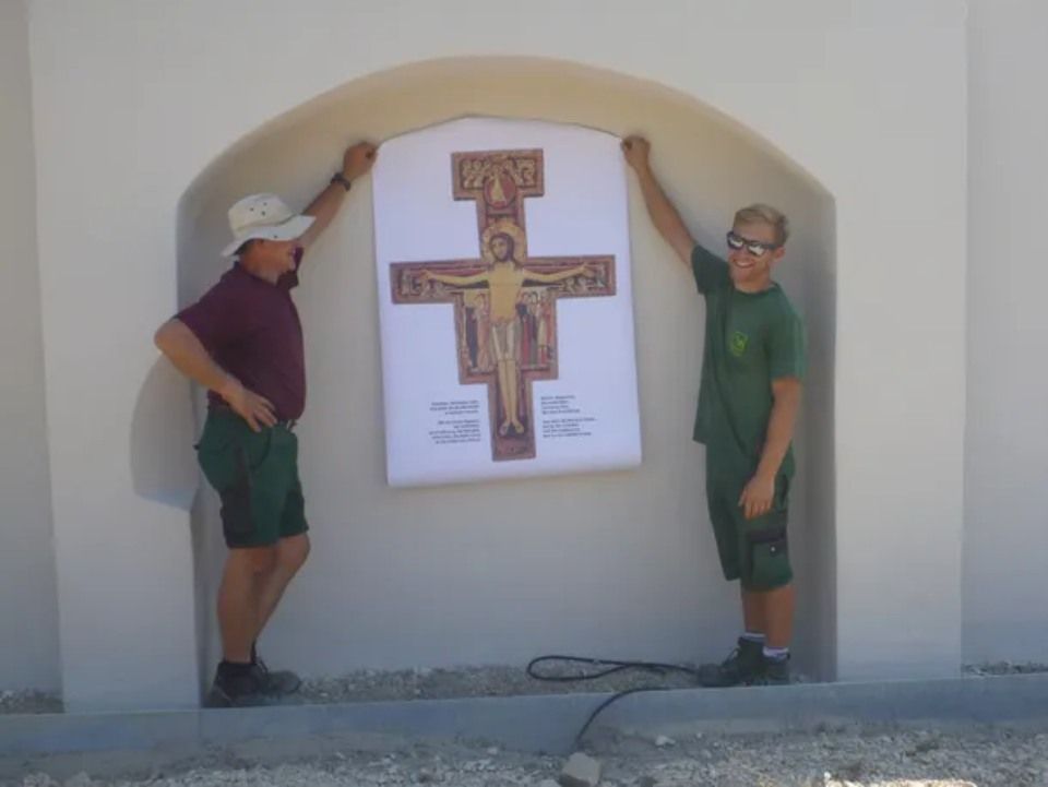 Zwei Männer halten ein großes Kruzifixplakat vor einer Bogennische in einem weißen Gebäude.