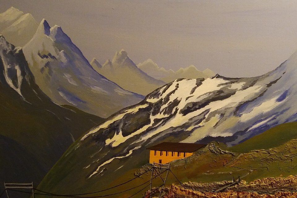 Berglandschaft mit einem Gebäude an einem Hang; schneebedeckte Gipfel im Hintergrund, bewölkter Himmel.