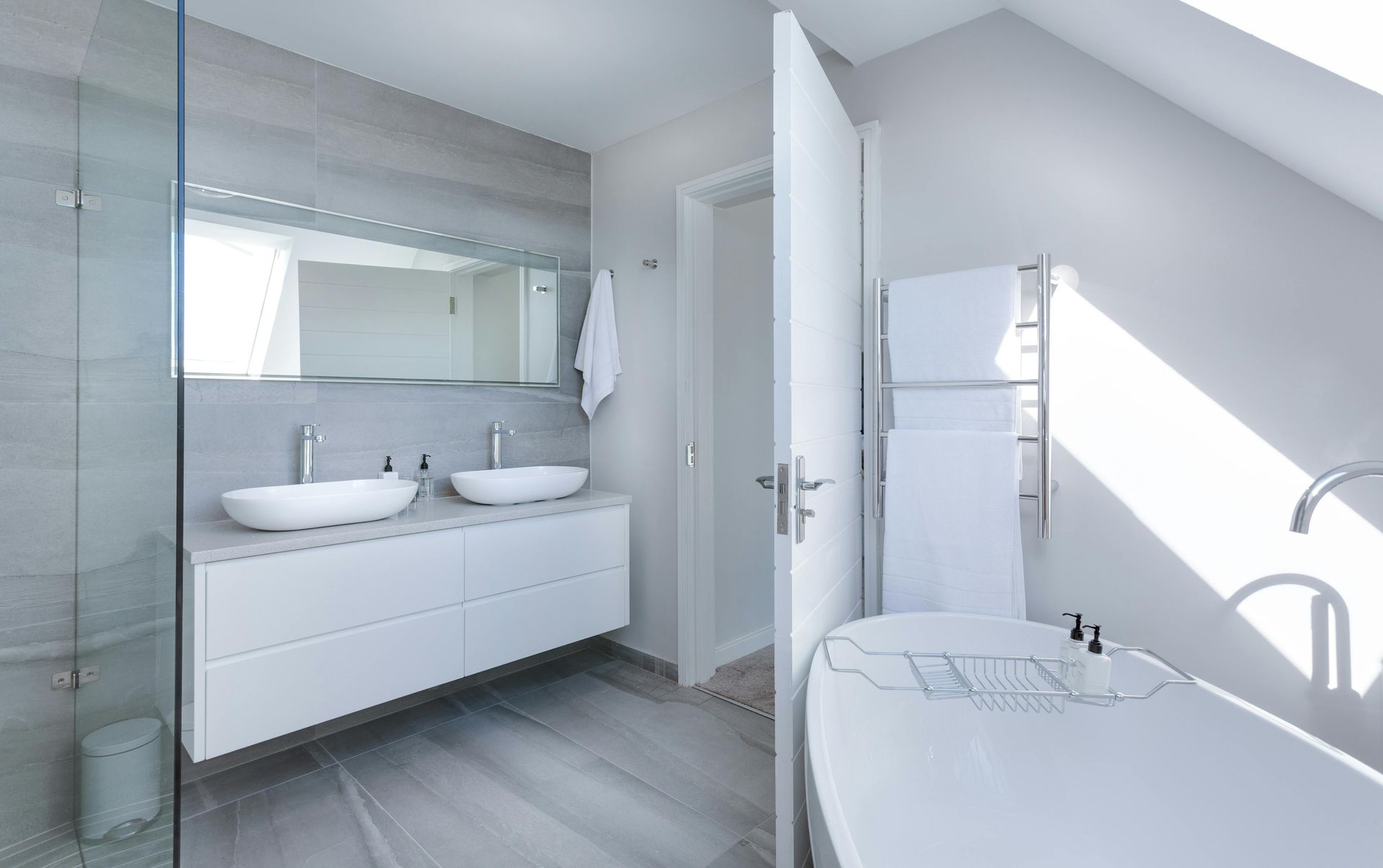 Modern white bathroom with a soaking tub, double vanity, and a heated towel rack. Sunlight streams through an open door.