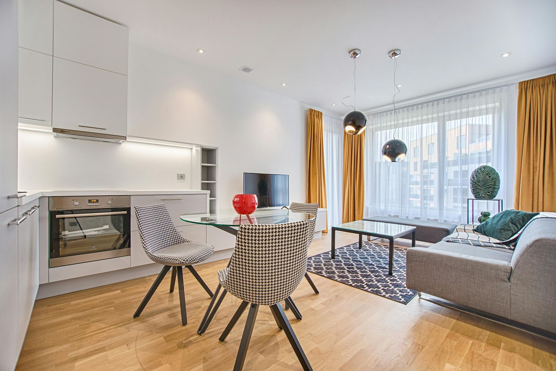 Modern apartment interior with a kitchen, dining area, and living room. Features white cabinetry, wooden floors, patterned chairs, and mustard-colored curtains.