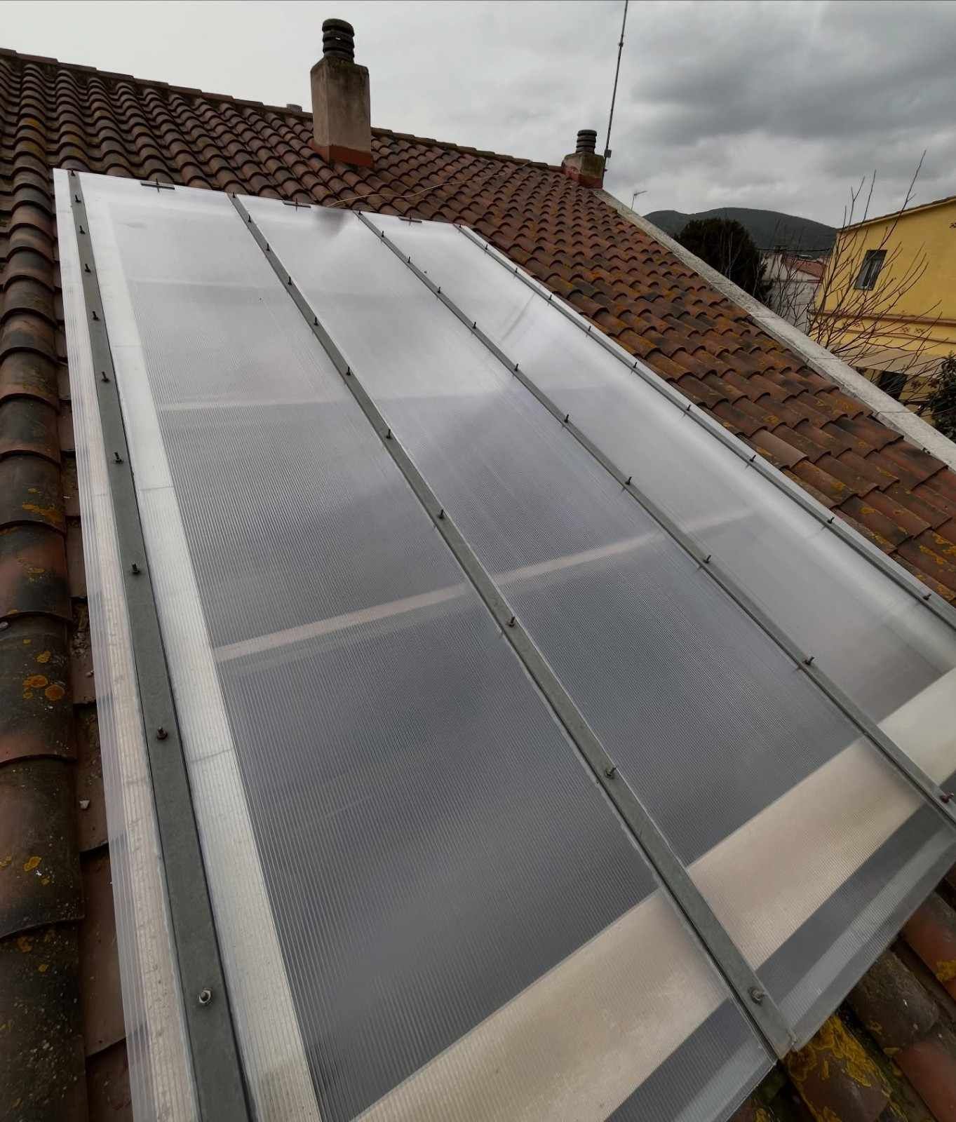 Un gran panel solar rectangular instalado en un tejado inclinado de tejas de terracota bajo un cielo nublado.