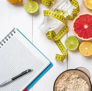 Cuaderno, bolígrafo, fruta, avena y cinta métrica sobre una mesa de madera blanca, sugiriendo alimentación saludable y pérdida de peso.