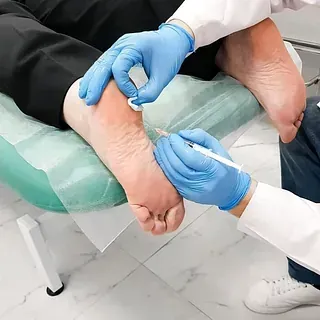 Un profesional médico inyecta el pie de una persona en un entorno clínico. El profesional usa guantes y bata de laboratorio.