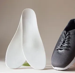 Par de plantillas ortopédicas blancas junto a una zapatilla deportiva gris sobre un fondo neutro.