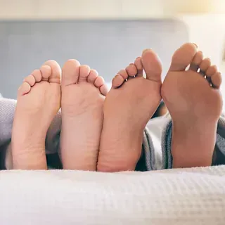Dos pares de pies descalzos relajándose en la cama bajo una manta blanca.