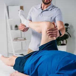 Fisioterapeuta examinando la pierna de un paciente en una sala iluminada. El paciente se recuesta en una camilla mientras el terapeuta le sujeta la pierna por la rodilla.