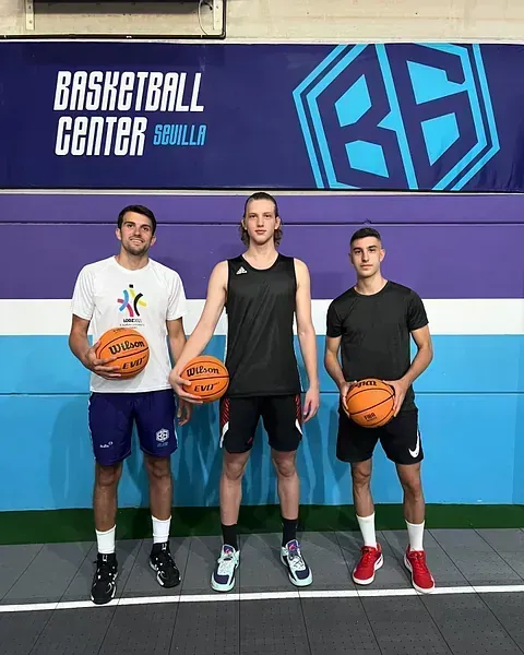 Tres jugadores de baloncesto se encuentran en un centro de baloncesto con balones Wilson. Llevan ropa deportiva y están frente a un fondo azul y morado.