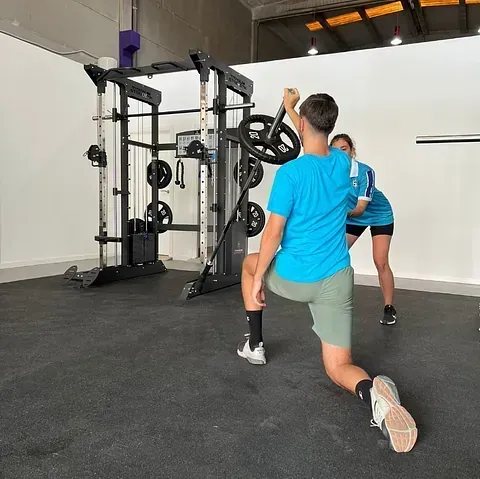 Un hombre realiza un ejercicio antiminas con una barra, asistido por una mujer en un gimnasio. El hombre está en posición de zancada, con camiseta azul, pantalones cortos y calcetines negros.