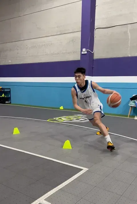 Jugador de baloncesto driblando el balón mientras se abre paso entre conos amarillos en una cancha gris. Viste un uniforme azul y blanco.