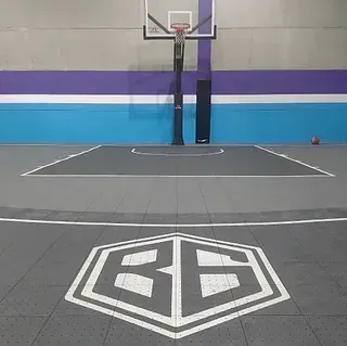 Cancha de baloncesto cubierta con suelo gris y verde azulado, pared morada y azul, y un logotipo en el centro. Un aro de baloncesto se ve al fondo.
