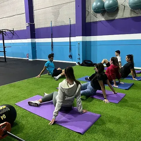 Clase de fitness grupal en un gimnasio, personas realizando ejercicios sentados sobre colchonetas, algunos mirando hacia adelante, otros hacia atrás, sobre césped artificial.