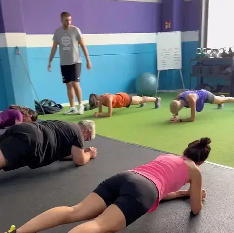 Una clase de fitness realiza planchas en el suelo del gimnasio. Un entrenador observa mientras la clase mantiene la postura.