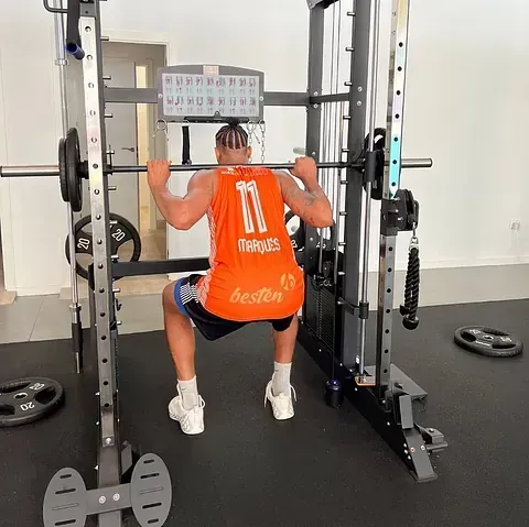 Una persona con camiseta naranja realiza una sentadilla en un gimnasio en casa. Usa un soporte de pesas y una barra.