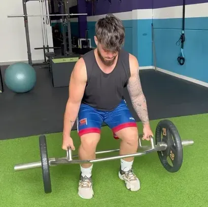Un hombre en un gimnasio realiza peso muerto con una barra hexagonal. Lleva una camiseta negra sin mangas, pantalones cortos azules y rojos, zapatillas deportivas y tiene tatuajes en el brazo.