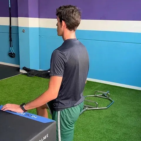 Un hombre en un gimnasio se apoya en una caja con la pierna derecha flexionada. Lleva pantalones cortos verdes y una camiseta oscura. El gimnasio tiene paredes azules y moradas.