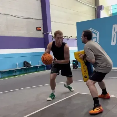 Un jugador de baloncesto dribla mientras mira a un entrenador que sostiene una rodillera, en un gimnasio con detalles en morado.