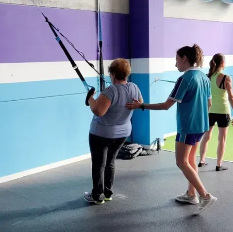 Mujer usando un entrenador de suspensión con la guía de un entrenador; otra mujer se ejercita al fondo. Gimnasio con paredes azules y moradas.