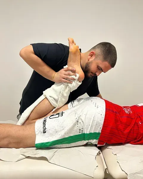 Un terapeuta estira la pierna de un paciente en una camilla, ayudándose con una toalla. El paciente está acostado, mientras el terapeuta se arrodilla, concentrado.