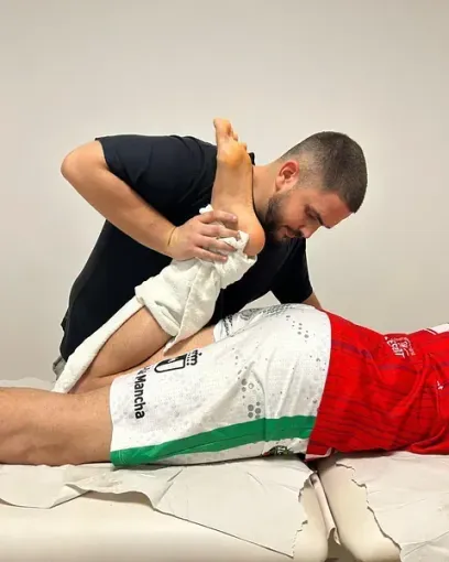 Un terapeuta estira la pierna de un paciente en una camilla, ayudándose con una toalla. El paciente está acostado, mientras el terapeuta se arrodilla, concentrado.