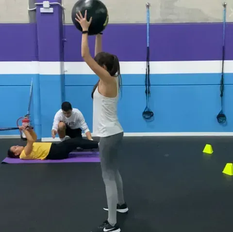 Mujer en el gimnasio, sosteniendo un balón medicinal. Dos personas al fondo hacen ejercicio mientras un entrenador observa.
