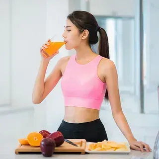 Una mujer con sujetador deportivo rosa bebe zumo y frutas en una tabla de cortar delante.