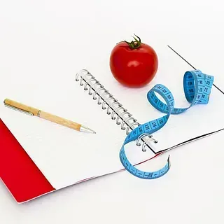 Un tomate rojo, una cinta métrica azul, un cuaderno abierto con bolígrafo, sobre una superficie blanca, sugieren salud o dieta.