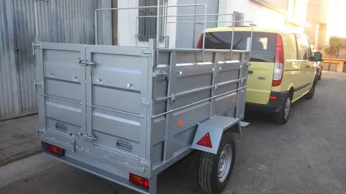Remolque utilitario gris enganchado a una furgoneta amarilla en una calle, con laterales metálicos y reflectores rojos
