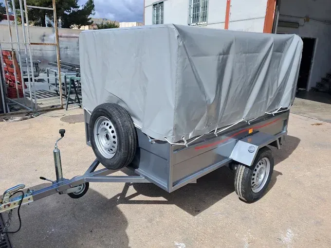Remolque utilitario cubierto con una lona gris, estacionado al aire libre junto a un edificio