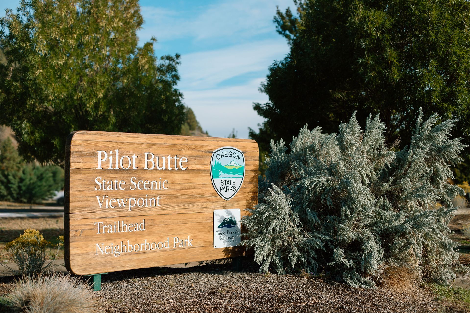 Sign for the Pilot Butte State Scenic Viewpoint Trailhead surrounded by juniper and sage bushes, with trees in the background under a clear sky.