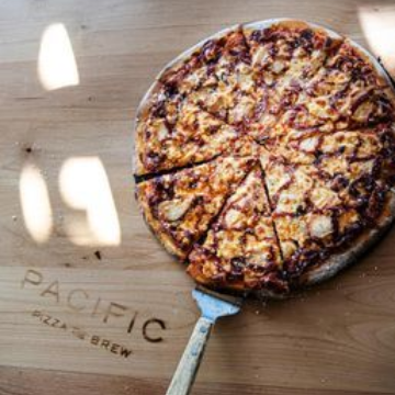 A barbecue pizza with chicken and bacon on a wooden cutting board branded with the Pacific Pizza & Brew logo, with a spatula lifting one slice, captured in bright, appealing lighting.