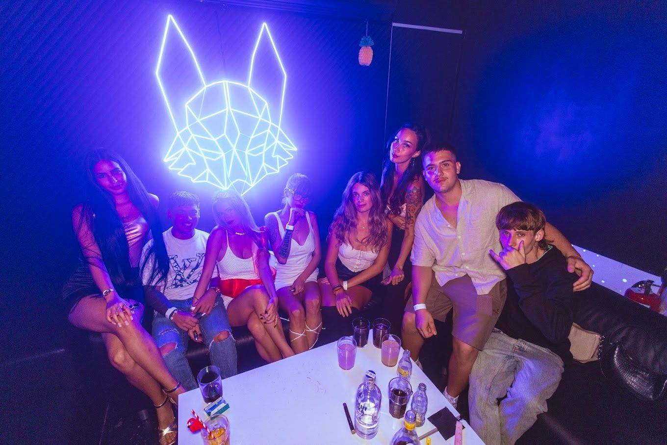 Grupo de personas en un club, iluminado por una cabeza de zorro de neón. Bebidas sobre una mesa, todos sonriendo.