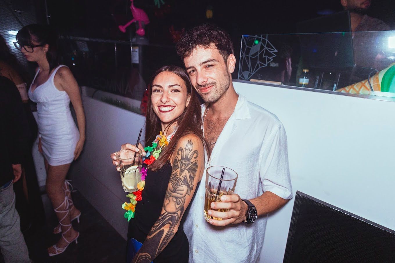 Una mujer con un tatuaje en el brazo y un hombre con camisa blanca sonríen en una discoteca.