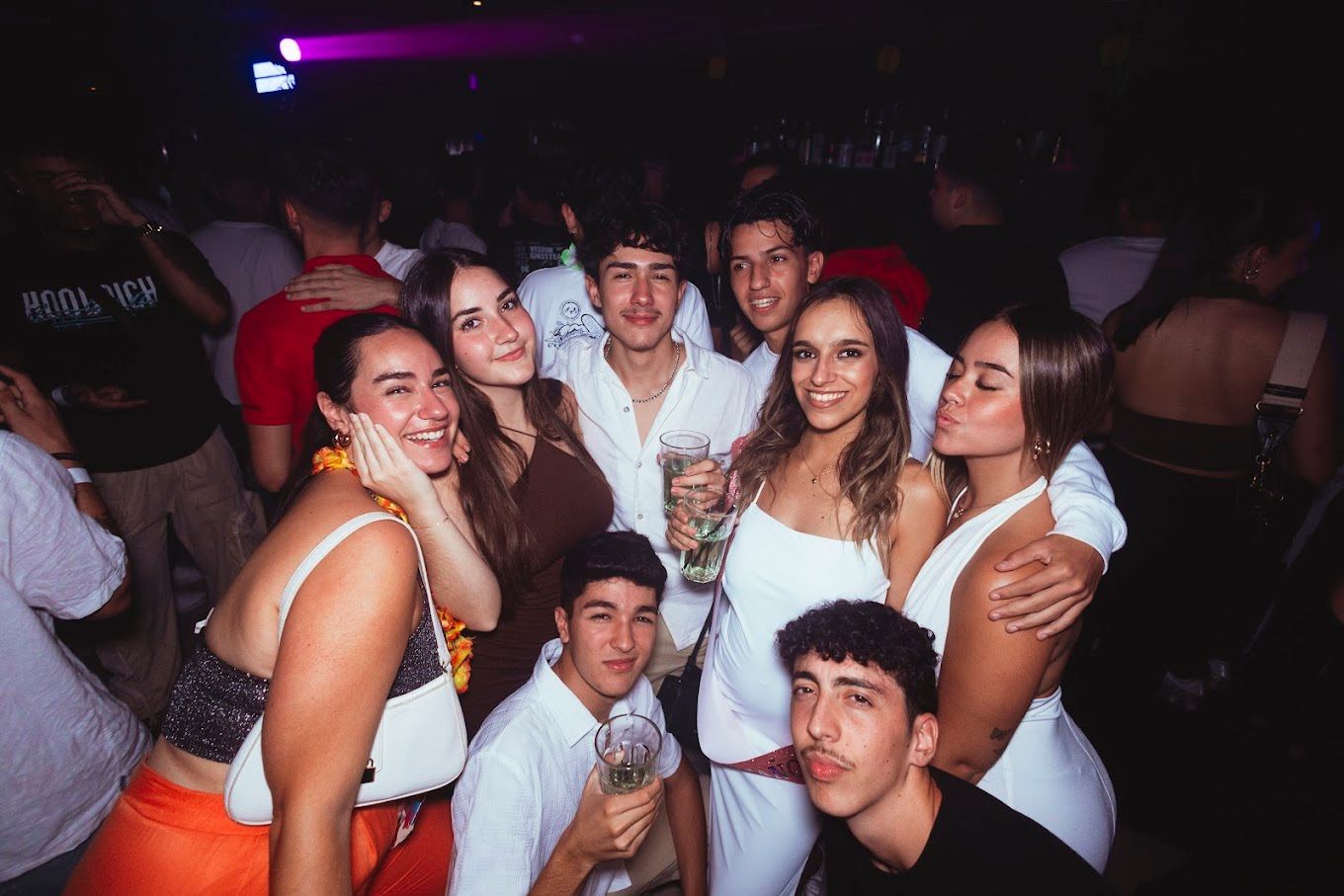 Grupo de personas sonriendo y posando en un club con poca luz. Vestimenta blanca y brillante; se ven las bebidas.